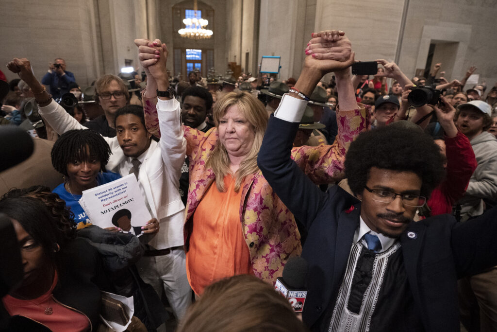 Image showing former Rep. Justin Jones, D-Nashville, Rep. Gloria Johnson, D-Knoxville, and former Rep. Justin Pearson, D-Memphis, holding hands with arms raised as they stand in the crowded Tennessee Capitol outside the House chamber.
