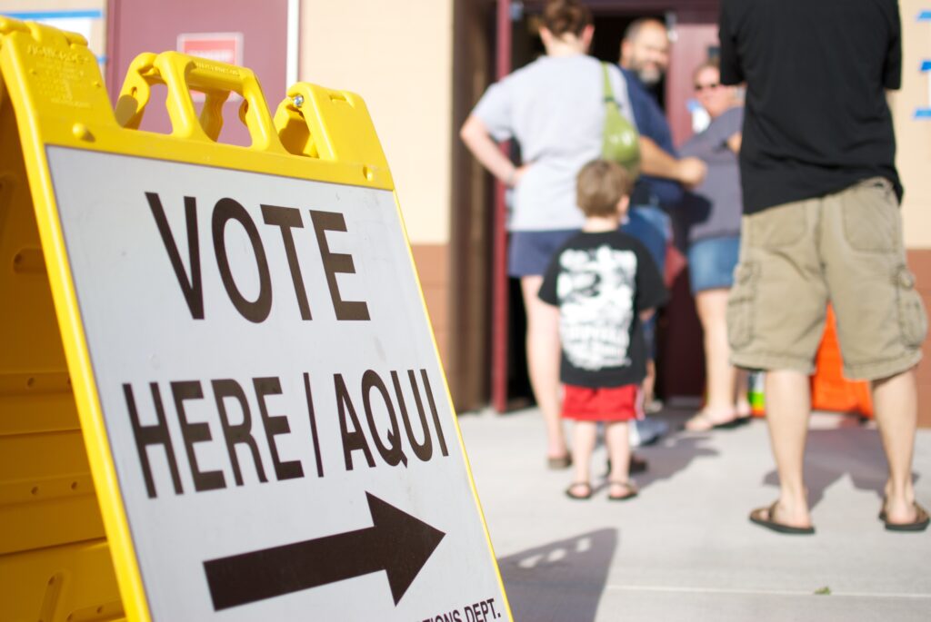 Yellow sign with VOTE HERE is standing by a line of people wating to get to the polls in Arizona