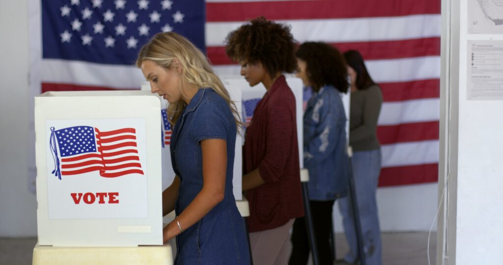 Four women filling in ballots and casting votes in booths at polling station, US flag on wall at back