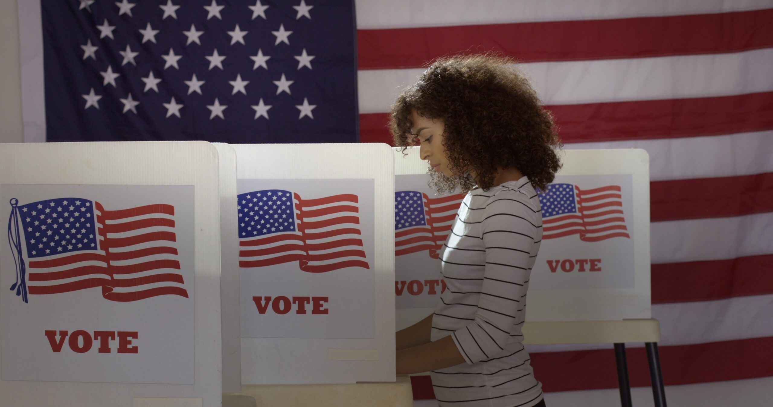 Profile medium shot young Hispanic woman-in polling station voting in a booth with US flag in background (Adobe Stock)