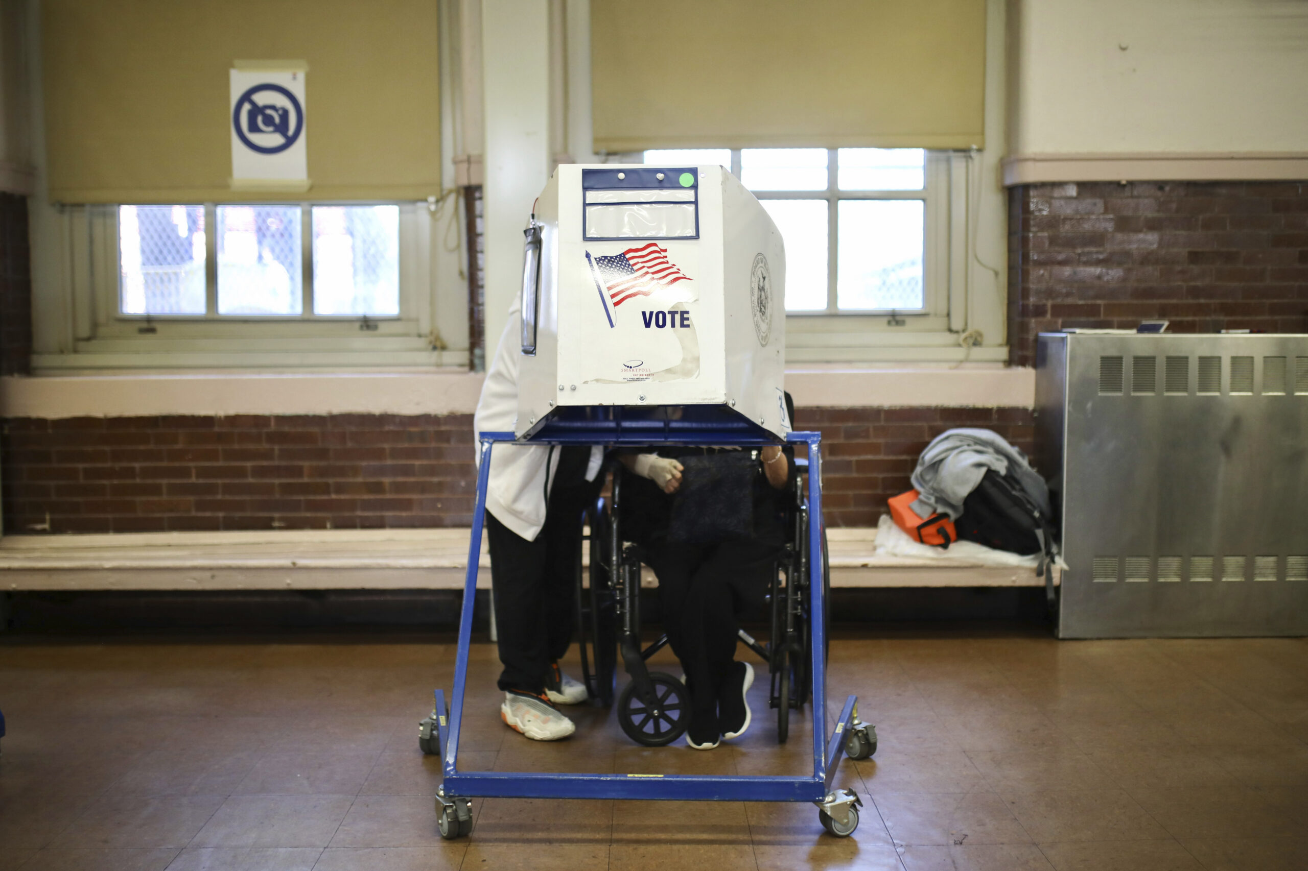 A person with a mobility device casts their ballot with the assistance of a poll worker, behind a panel reading with an American flag and the word "vote."