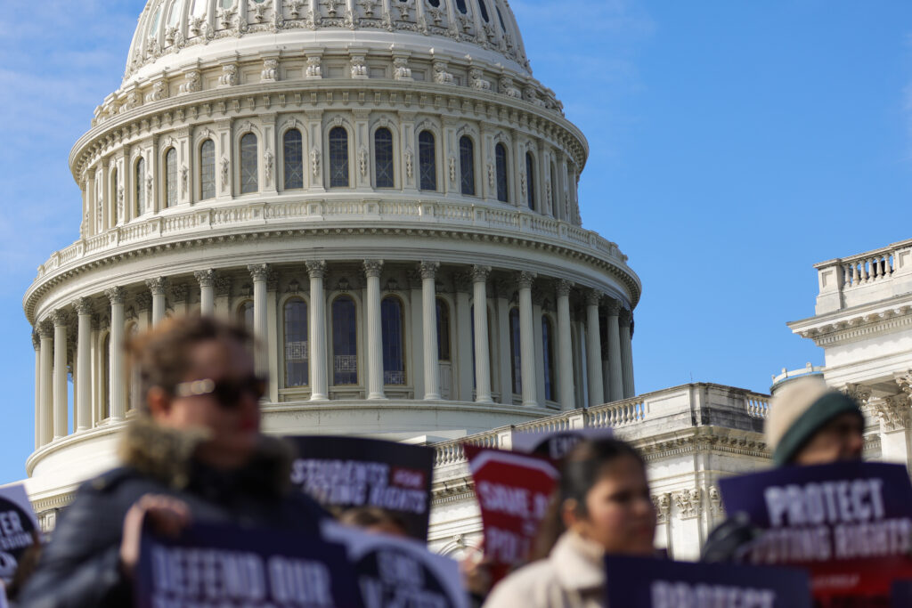The dome of the U.S. Capitol building is seen beyond people holding signs at a rally and press conference against the SAVE America Act at the U.S. Capitol in Washington, D.C. on March 18, 2026. The bill being debated in the Senate requires voters to provide proof of citizenship and a photo ID in order to vote in U.S. elections. (Photo by Bryan Dozier/NurPhoto via AP)